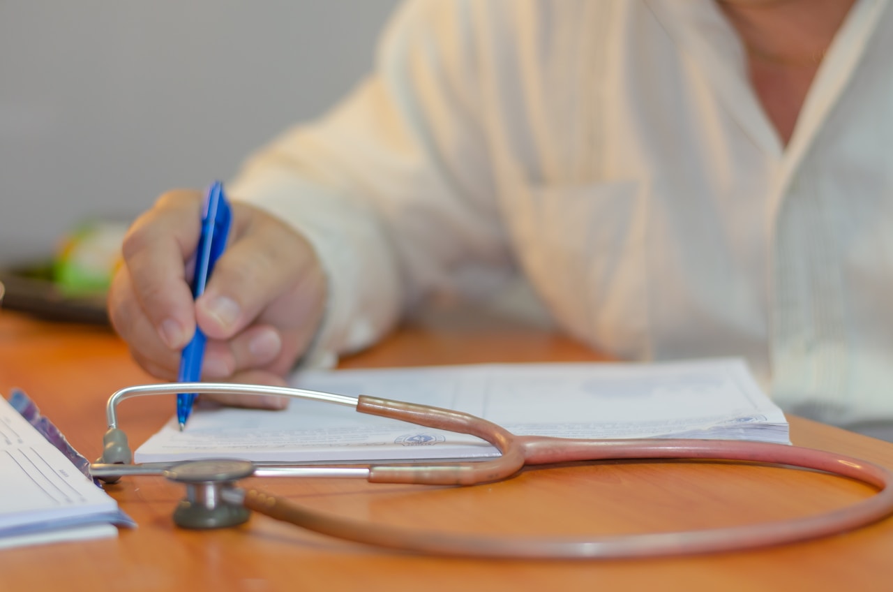 A doctor writing on a piece of paper on a desk with a stethoscope